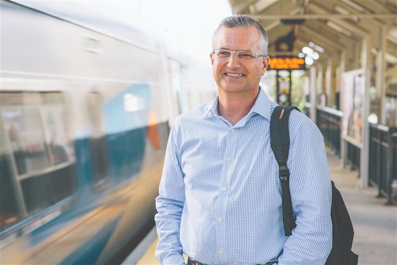 A man is standing on a train platform. A train is moving at a blur to his left. He's in the right foreground, wearing a button-up collar shirt, with a black backpack hanging over this left shoulder. He's wearing glasses and smiling. He has short hair, white at the temples and graying on top.