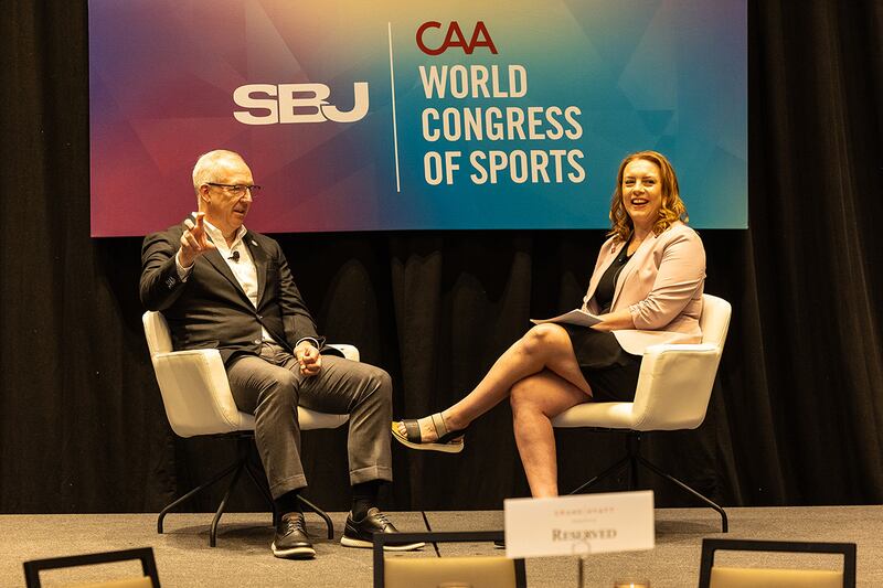 A man with graying hair in a brown jacket, white shirt and light brown pants with an SEC button, Greg Sankey, sits across on a stage from a woman with red hair, a pink jacket, black skirt, SBJ's Rachel Axon.